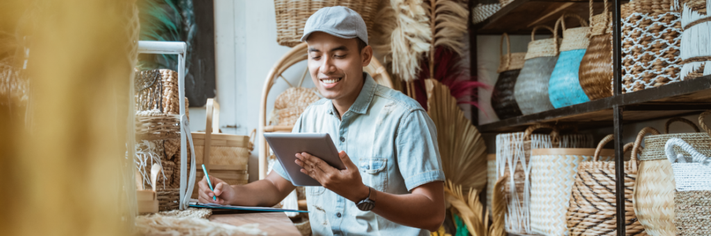 A man in a cap and denim shirt uses a tablet and takes notes in a craft store surrounded by baskets.