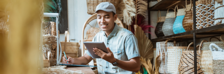 A man in a cap and denim shirt uses a tablet and takes notes in a craft store surrounded by baskets.
