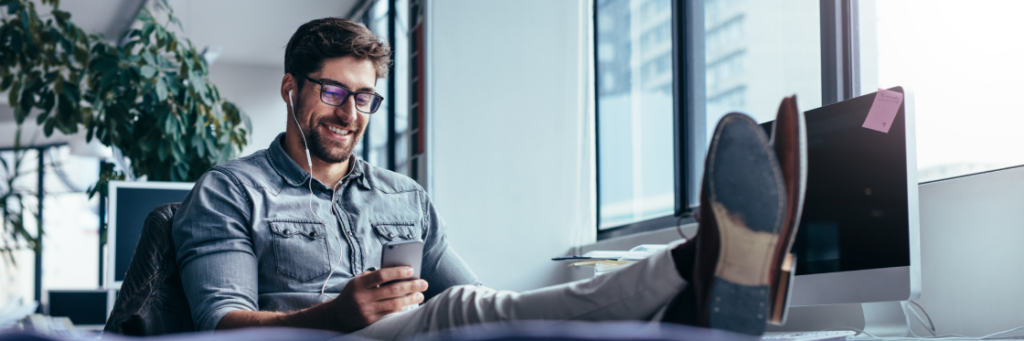 Man in casual attire using a smartphone with feet up on a desk in an office setting.