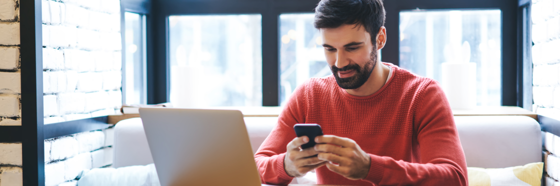 YouTube TrueView video advertising campaigns Man in red sweater using smartphone with laptop on table in a modern room with large windows.
