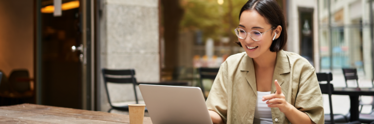 A smiling woman wearing glasses using a Biteable video maker on her laptop at an outdoor cafe table.