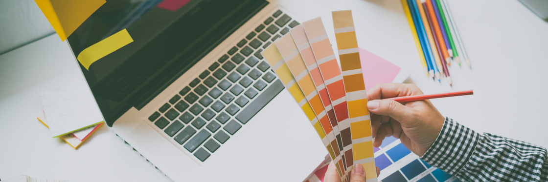 Color theory basics for video makers Person holding color swatches with a pencil in hand beside a laptop and colored pencils on a desk.