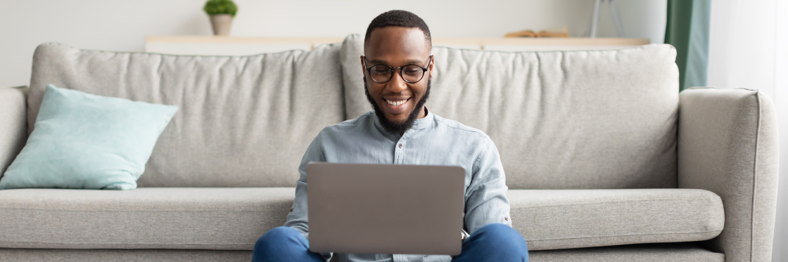 A smiling man sitting on the floor with a laptop on his lap.