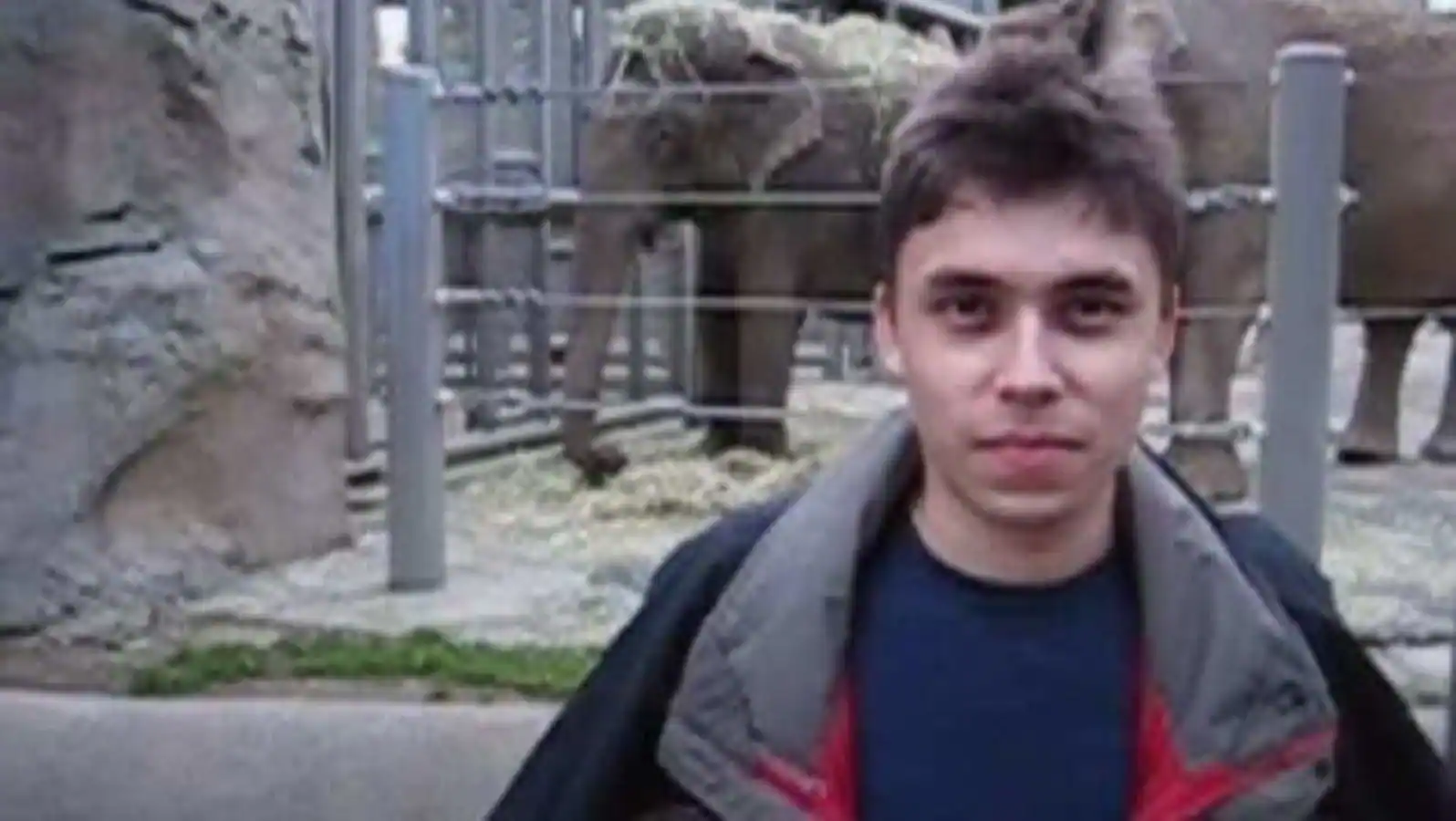 A young man with tousled hair and a jacket stands in front of an elephant enclosure, creating a scene perfect for a Biteable video maker project with visible elephants in the background.