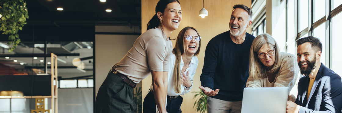 Five colleagues sharing a light-hearted moment while looking at a laptop in an office environment.