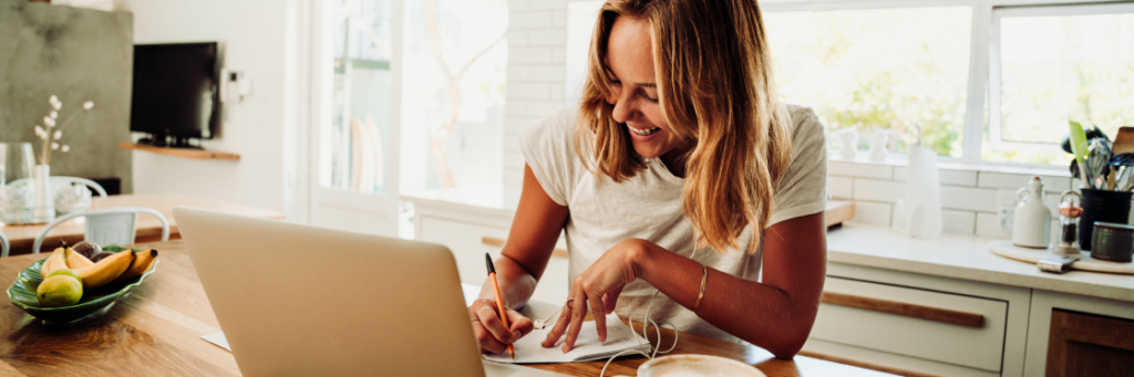 A smiling woman writing in a notebook while using a laptop at a kitchen table.