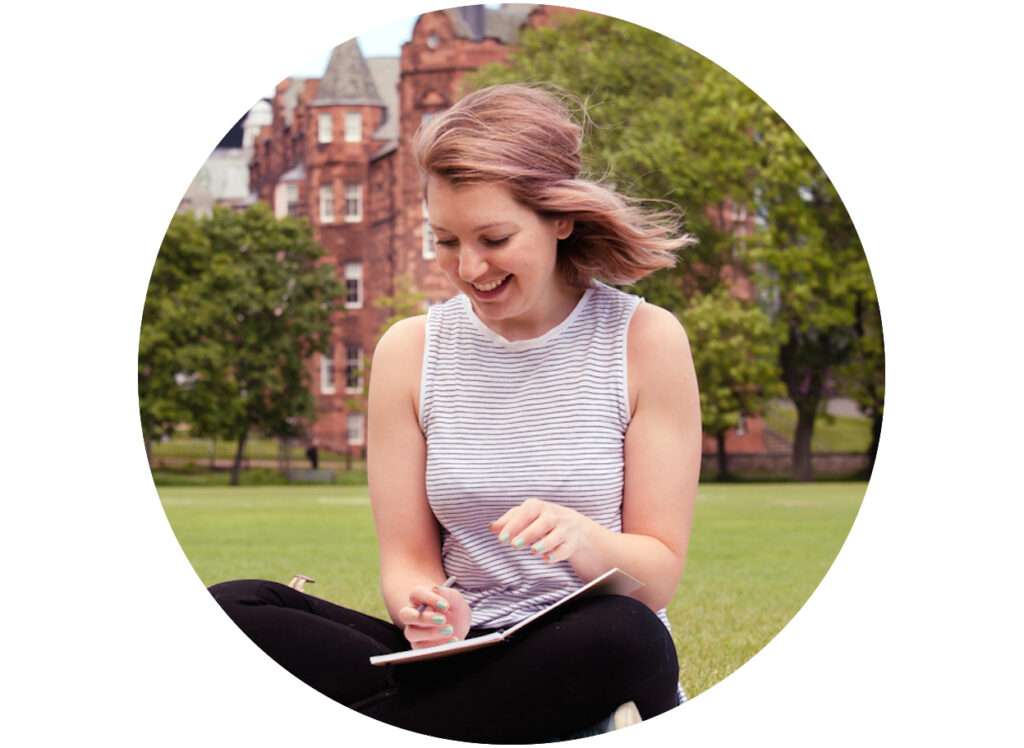 A young woman smiling and looking down at her tablet while sitting on the grass in front of a brick building.