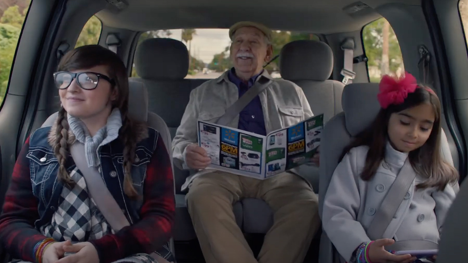An elderly man reads a magazine between two young girls, one engrossed in a tablet and the other wearing headphones, in a Biteable video maker-themed car.