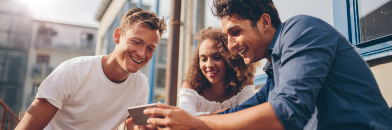 Three friends smiling and looking at a smartphone together outdoors.