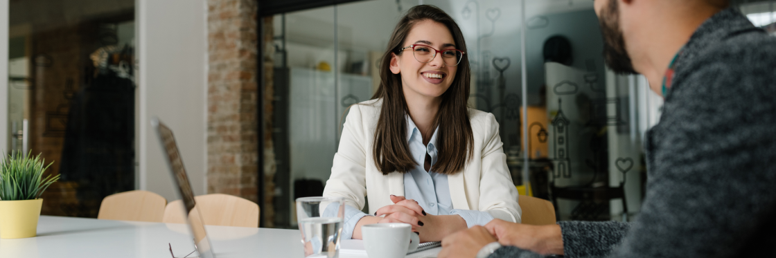 Outstanding video resume examples to get you hired Woman in glasses and white blazer smiling during a meeting with a male colleague in a modern office setting.