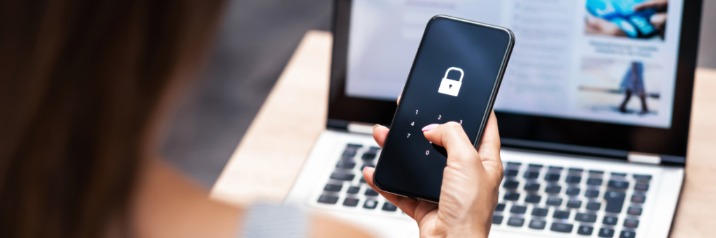 Person entering a security passcode on a smartphone with a laptop in the background.