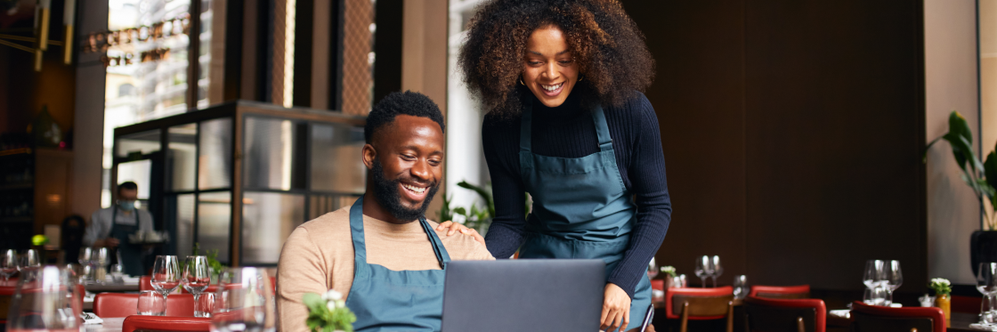 40 sizzling restaurant marketing ideas to get you more traffic A man and a woman in aprons smiling at a laptop screen while using Biteable video maker in a modern restaurant setting.
