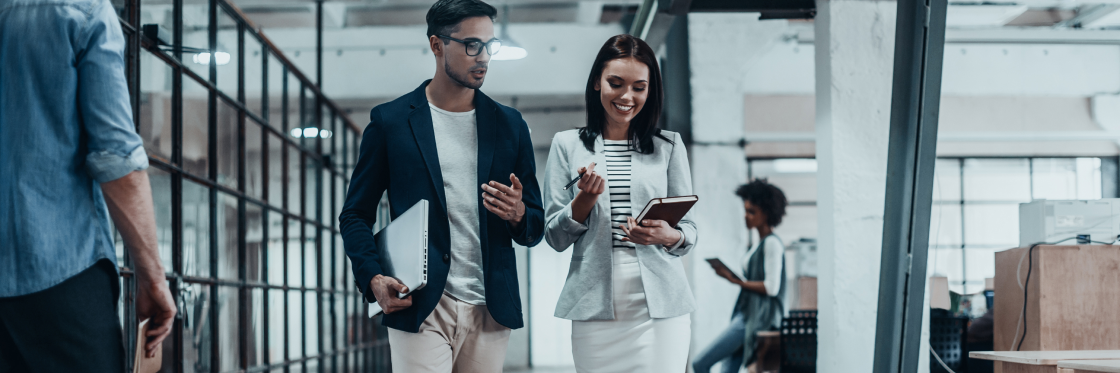 Two professionals walking and discussing in a busy office setting, with a focus on a man and a woman who are looking at a digital tablet featuring the Biteable video maker.
