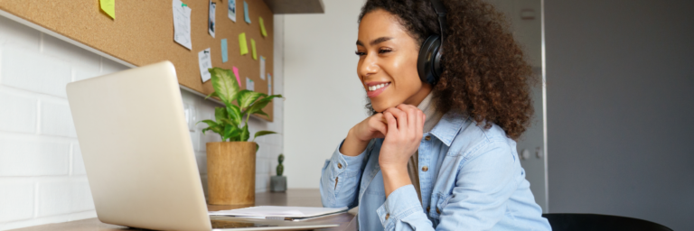 A smiling woman with headphones using a laptop at a desk.