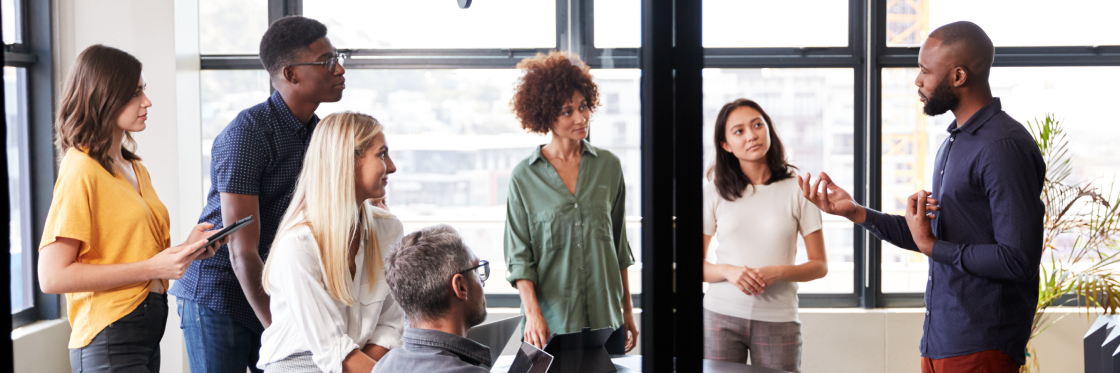 A diverse group of professionals engaged in a discussion with one individual presenting to the others in a bright office space using Biteable video maker.