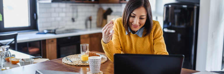 Woman in yellow sweater eating breakfast and working on laptop in kitchen.