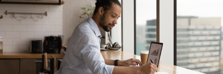 A man in business casual attire working on a tablet at a kitchen counter with a coffee cup beside him.