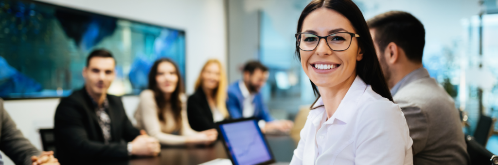 A smiling woman in a white shirt at the forefront of a business meeting with colleagues in the background, engaging with Biteable video maker.