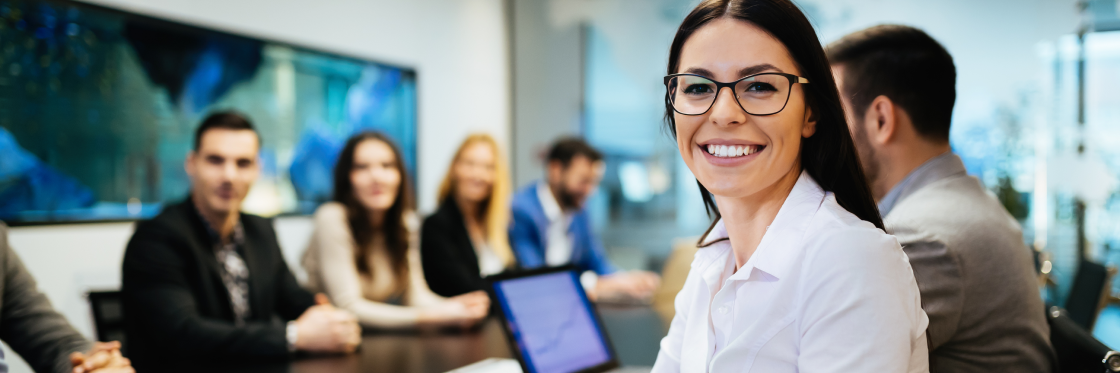 A smiling woman in a white shirt at the forefront of a business meeting with colleagues in the background, engaging with Biteable video maker.