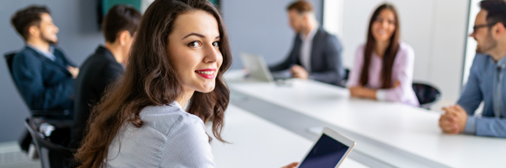 Woman with tablet smiling in a meeting room with colleagues in the background.