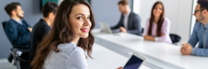 Woman with tablet smiling in a meeting room with colleagues in the background.