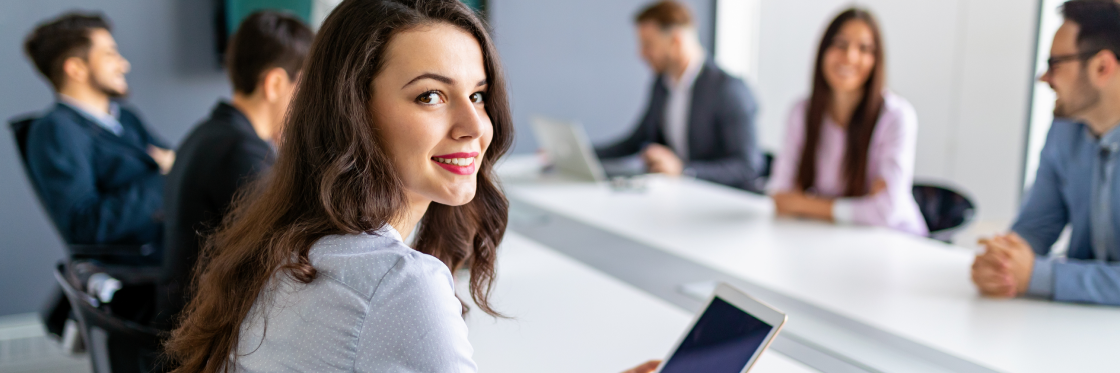 Woman with tablet smiling in a meeting room with colleagues in the background.