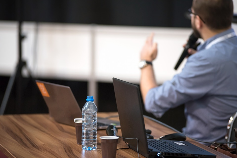 Presenter gesturing during a conference with laptops and a water bottle on the table.