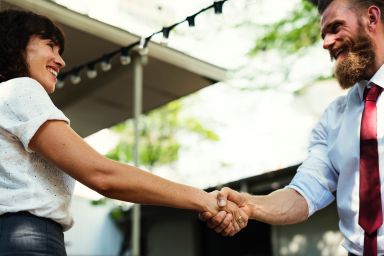Two people shaking hands outdoors with smiles on their faces.
