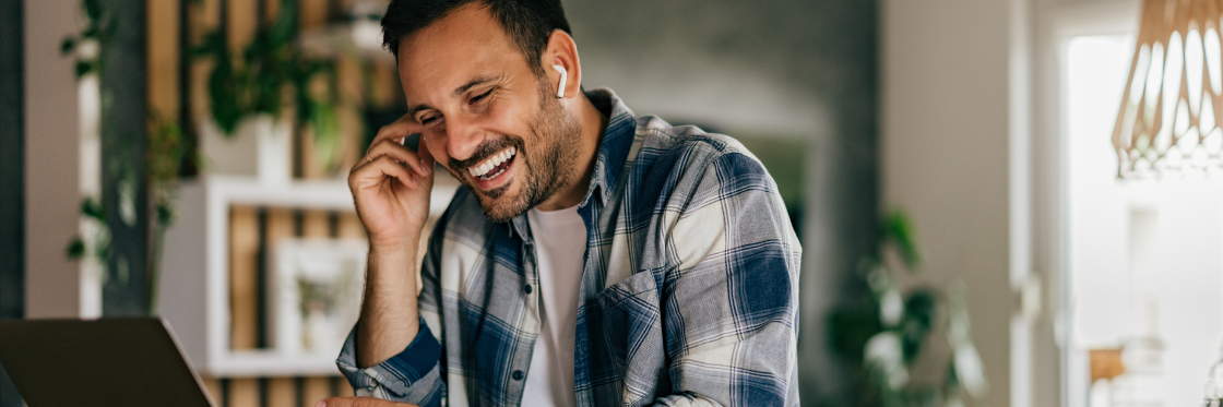 A man in a plaid shirt laughs while talking on the phone, sitting at a desk in a room decorated with plants and video testimonials.