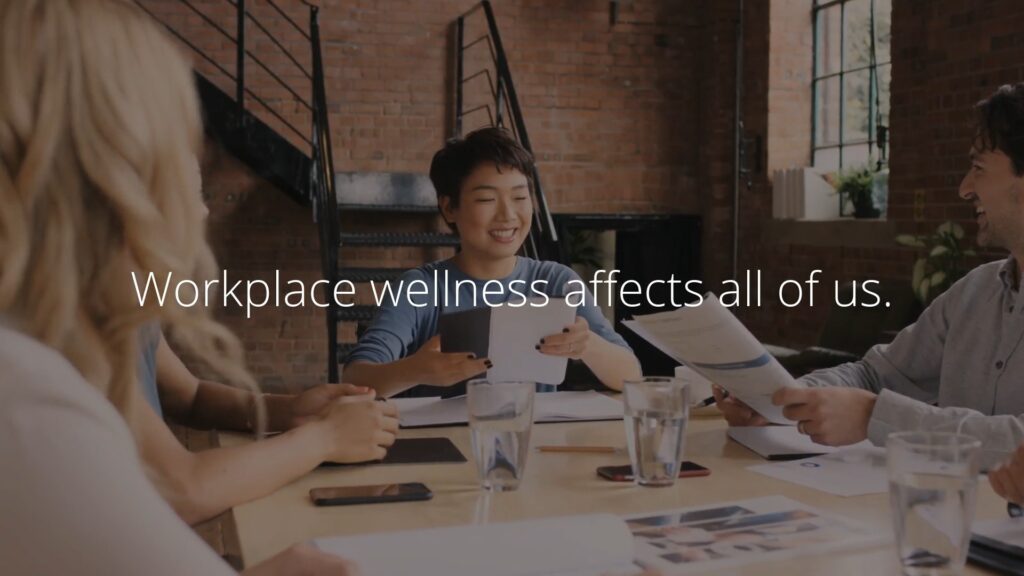 Group of professionals discussing documents at a meeting table with a Biteable video maker presentation in the background displaying the message, "workplace wellness affects all of us.