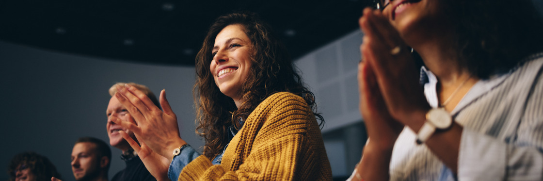 Event marketing ideas to inspire your brand Diverse group of people clapping enthusiastically in an auditorium, focusing on a smiling woman in a yellow sweater featured in a Biteable video.