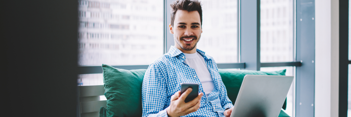 Facebook Marketing Man in a blue plaid shirt sitting with a laptop on his lap and holding a smartphone, smiling in a modern office with large windows, using the Biteable video maker.