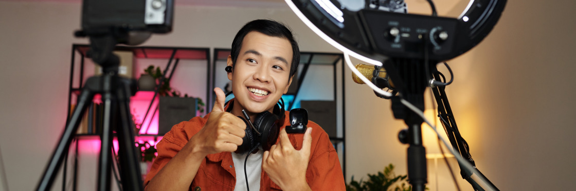 The best video lighting kits to make your videos look pro Young man holding a Biteable video maker camera, giving a thumbs up between two ring lights and tripods, with a pleasant expression in a well-lit room.