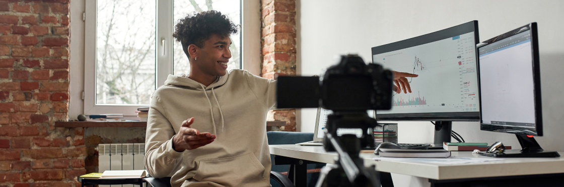 The best vlogging camera 2021 A young man records a video in a brick-walled office, gesturing towards graphs on his computer screen, with a Biteable video maker camera in the foreground.