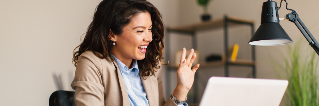 The ultimate guide to connecting with customers via video A woman in a beige blazer smiling and gesturing during a video call on her laptop in a home office, using Biteable video maker.