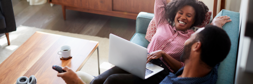 A woman lies on a couch using a laptop to explore Biteable video maker, facing a man who holds a remote control, in a cozy living room setting.