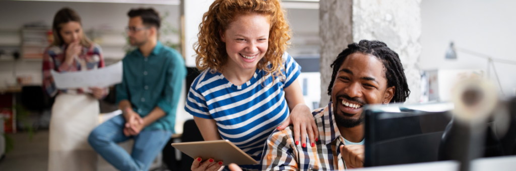 Two colleagues, a smiling man and woman, working together on a Biteable video maker project using a tablet in a busy office setting.