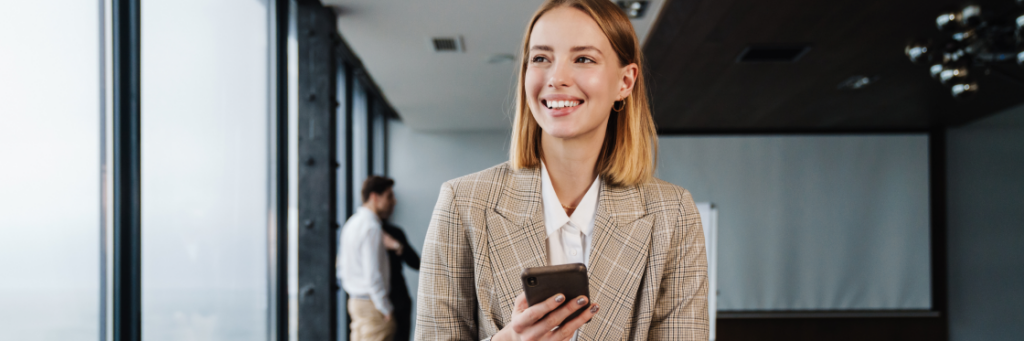 A smiling woman in a plaid blazer using the Biteable video maker on a smartphone in a modern office setting, with a male colleague in the background.
