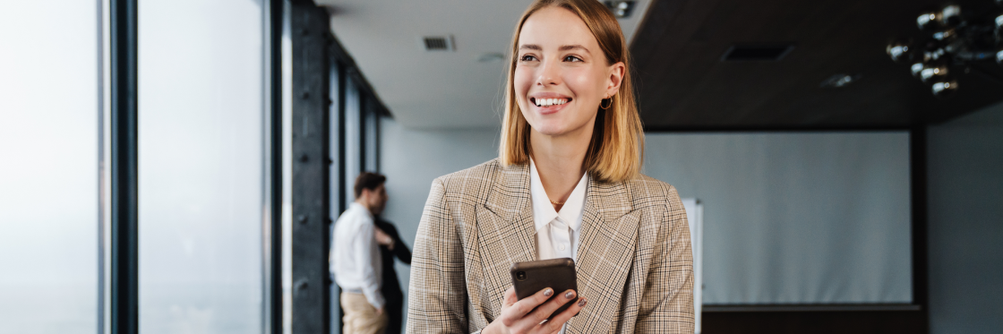A smiling woman in a plaid blazer using the Biteable video maker on a smartphone in a modern office setting, with a male colleague in the background.