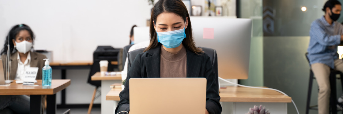 Return to office How to get it right with video Woman wearing a mask works on a laptop using Biteable video maker in a socially distanced office setting with colleagues in the background.
