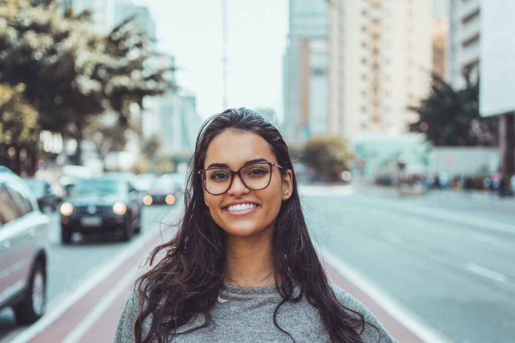 A smiling young woman with glasses standing on a city street, aiming to motivate Gen Z, with blurred traffic and buildings in the background.