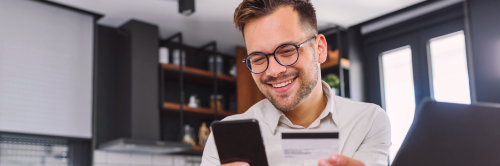 Smiling man with glasses using smartphone and laptop in a modern kitchen.