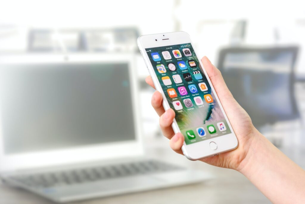 A person holding an iPhone with various app icons visible on the screen, with a blurred office background featuring a laptop.