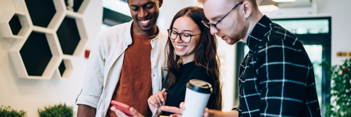 How brands use social media content to grow their business Three colleagues, one Black man and two white individuals, a woman and a man, look at a smartphone and smile together in a modern office setting.