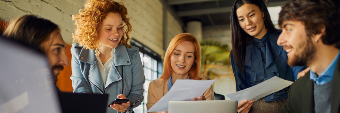 How to speed up your team’s content creation process without compromising on quality Five young professionals discussing work documents and a laptop in a lively, modern office setting.