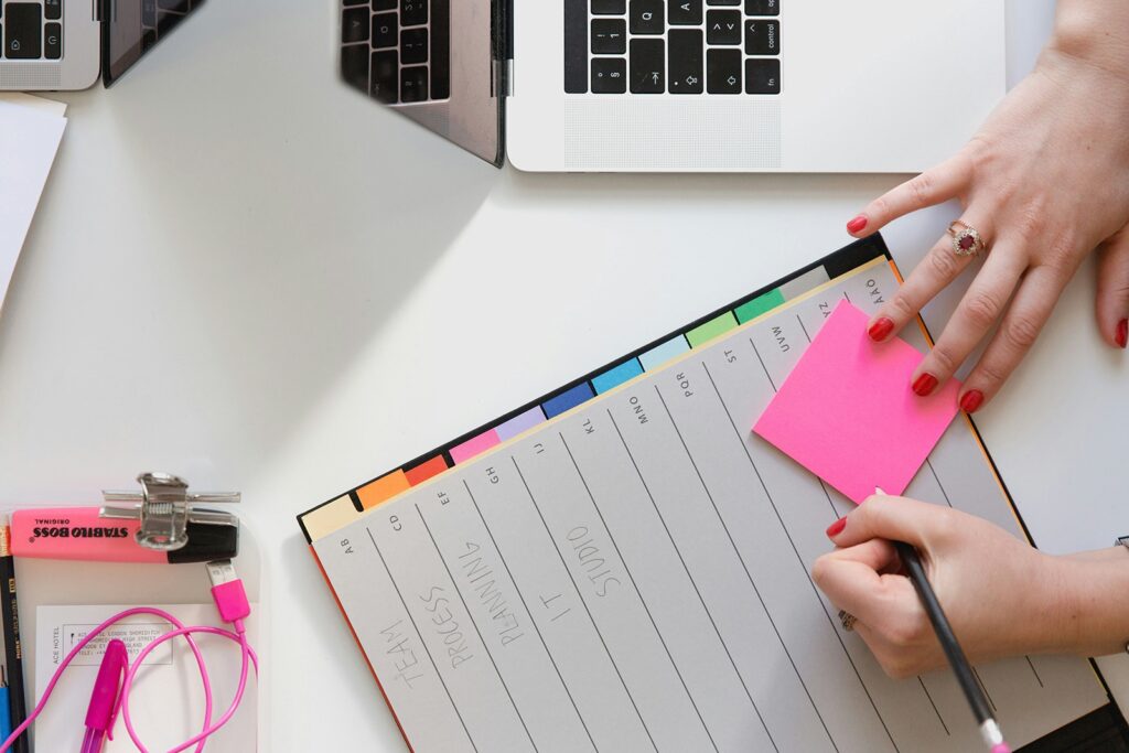 Hands holding a pink sticky note and pencil over a planner with colorful tabs and labeled sections, surrounded by two open laptops, earphones, and a highlighter.