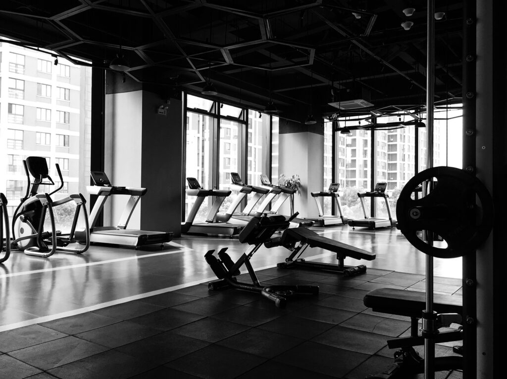 A gym with several treadmills, weightlifting equipment, and benches, set against large windows overlooking an urban landscape. Black and white photo.