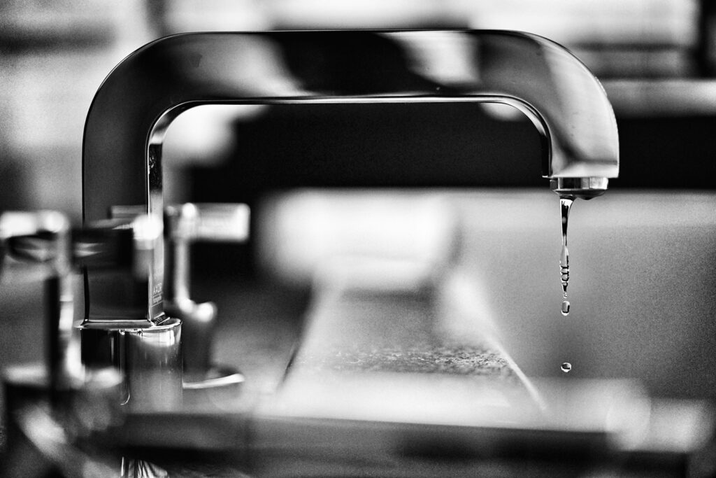 Close-up of a modern faucet with a droplet of water falling from the spout, captured in black and white.