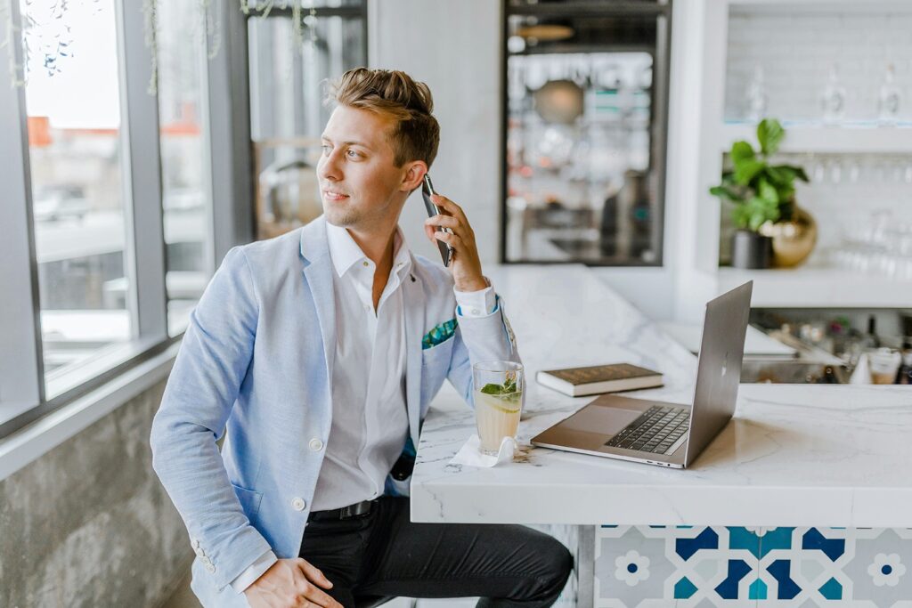 Person in a light blue blazer sitting at a bar counter talking on a phone, with a laptop and a drink nearby.