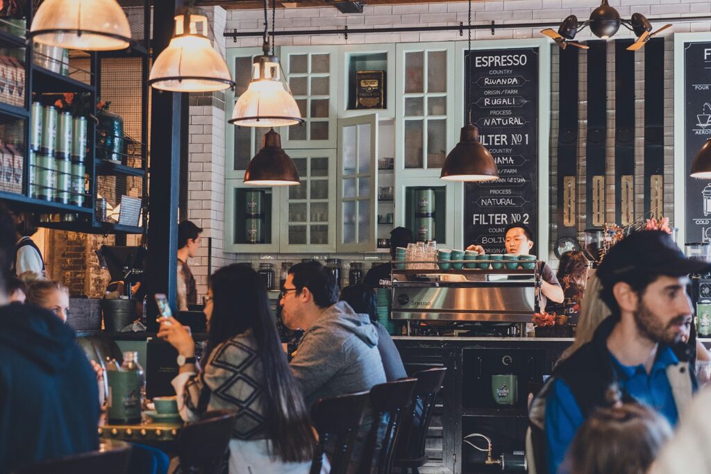 People sitting and chatting in a busy coffee shop, with a barista preparing drinks behind the counter.